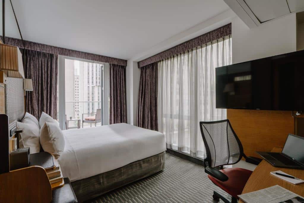 No quarto do The Jewel Hotel há uma cama de casal ao centro, com cabeceira de madeira. À esquerda, uma secretária com cadeira preta e uma TV na parede; à direita, uma poltrona junto à janela com cortinados escuros.