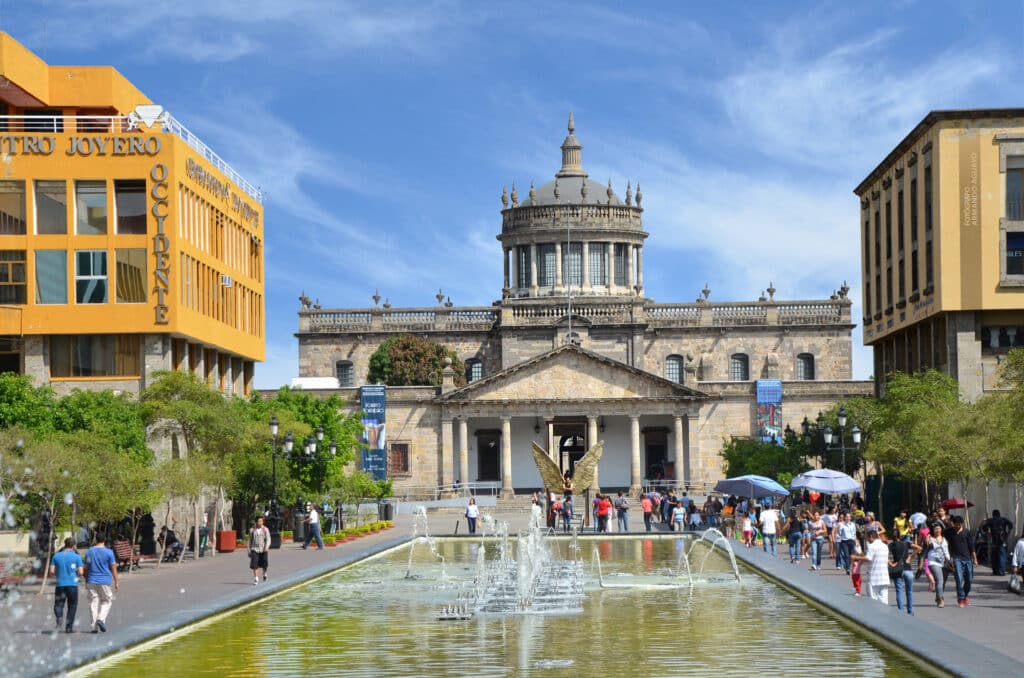 Edifício histórico magnífico com cúpula elevada, numa praça com fonte e movimentado centro urbano com pessoas a circular; trata-se do Hospício Cabañas.