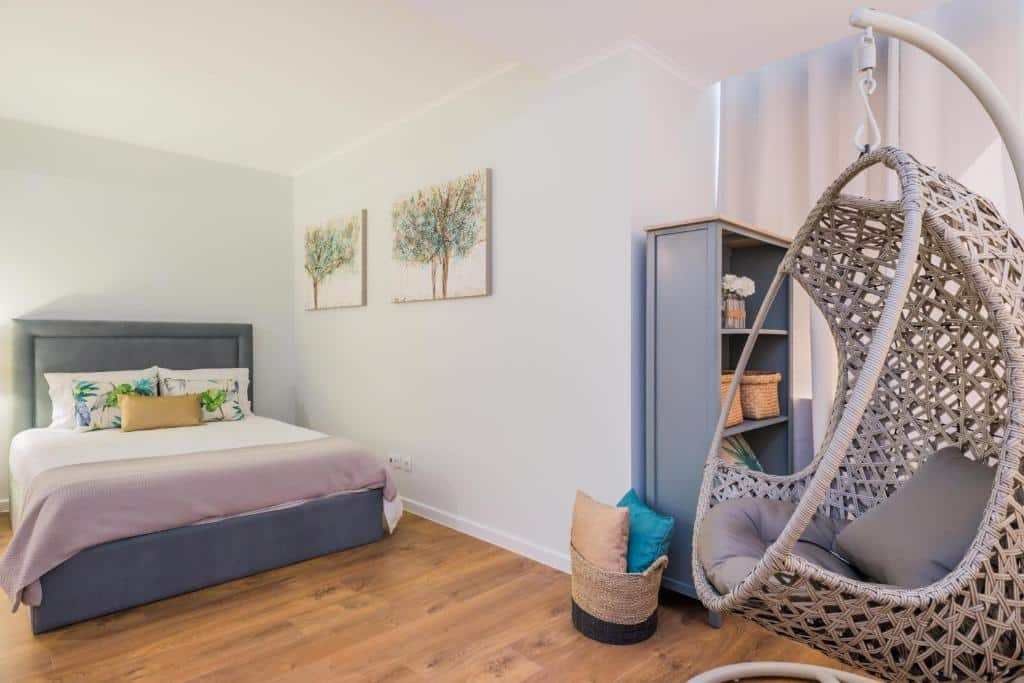Quarto do 137 LuxResidence com uma cama de casal com almofadas, dois quadros com flores, uma estante com quatro prateleiras e uma cadeira suspensa com almofadas