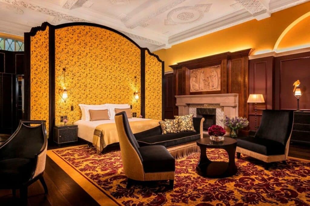 Quarto do L’oscar London com uma cabeceira alta amarela com desenhos de flores pretas, cama de casal, uma lareira, duas poltronas estofadas pretas e um amplo tapete florido em vermelho e dourado.