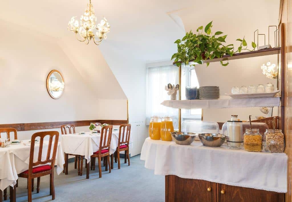 Sala de refeições bem iluminada no Pension Dr. Geissler, com mesas postas e uma zona de buffet com sumo, cereais, taças e pratos.
