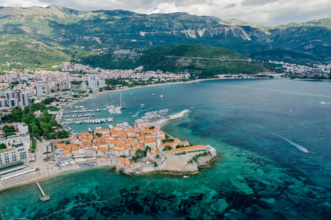 Vista aérea da cidade costeira de Budva, no Montenegro, com edifícios de telhados alaranjados, rodeada por uma marina e por colinas verdes sob um céu parcialmente nublado.