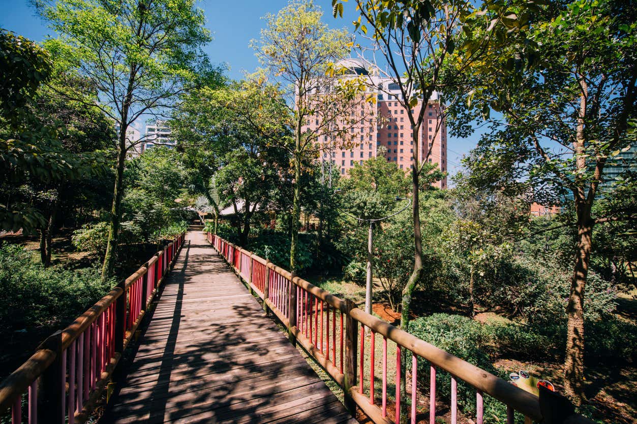 Passadiço de madeira com guardas vermelhas no Parque La Presidenta, atravessando um jardim verde em direção a edifícios altos, sob um céu azul.