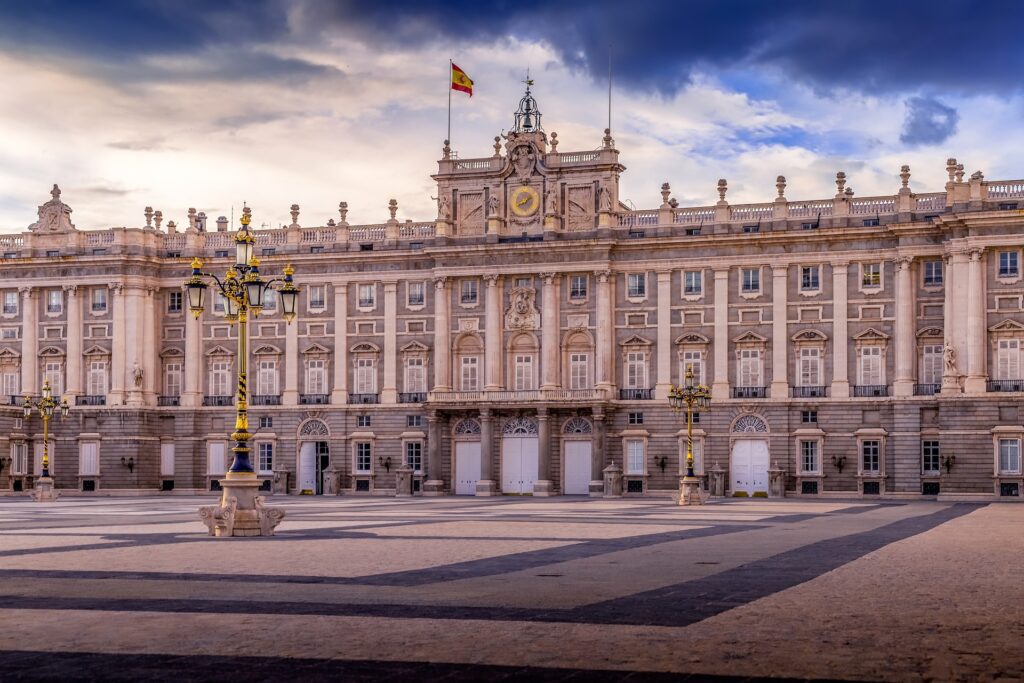 Palácio bege repleto de colunas e janelas. Há candeeiros na praça em frente, e uma bandeira de Espanha tremula ao vento perto do relógio que fica no centro do edifício.