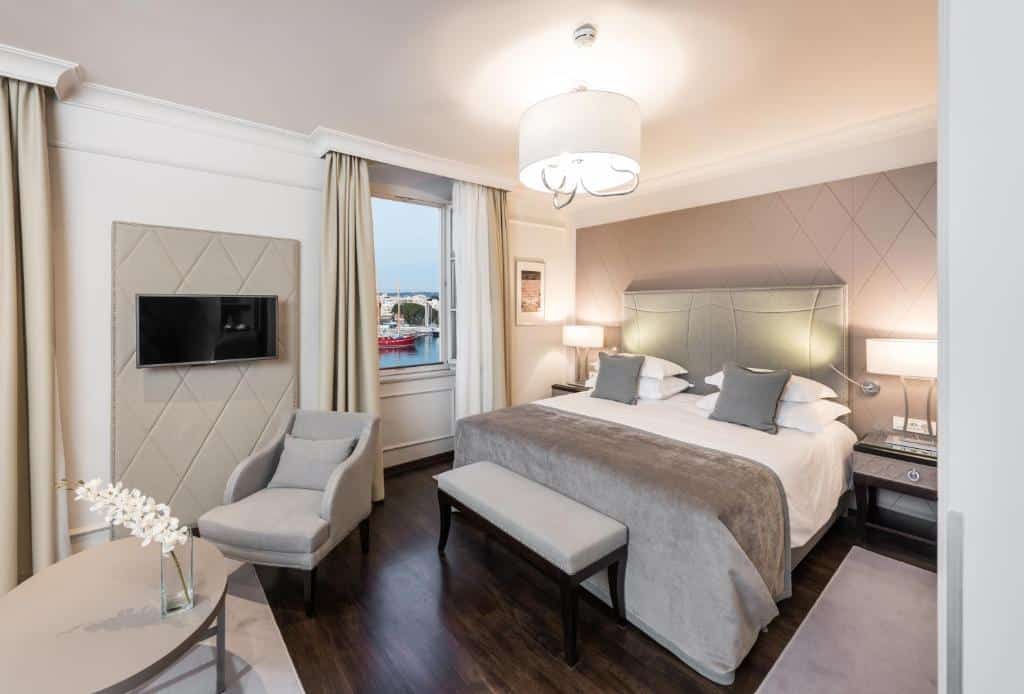 Quarto de hotel luxuoso com cama de casal, poltrona cinzenta e vista para marina com barcos, no Bastion Heritage Hotel, na Cidade Velha