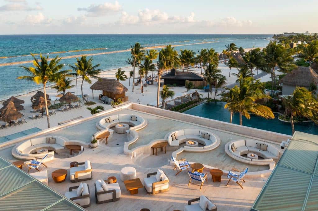 Fairmont Mayakoba com vista para praia de areia branca e mar azul-turquesa. A foto mostra uma área de lazer do hotel com várias poltronas e sofás. Mais abaixo, há árvores e espreguiçadeiras na areia.