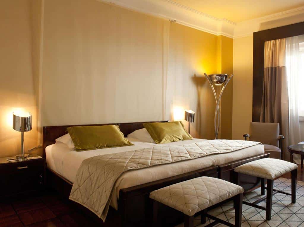 Quarto do Hotel Britania com cama grande, duas almofadas verdes, dois bancos, candeeiros modernos, uma poltrona e uma janela com cortinas.