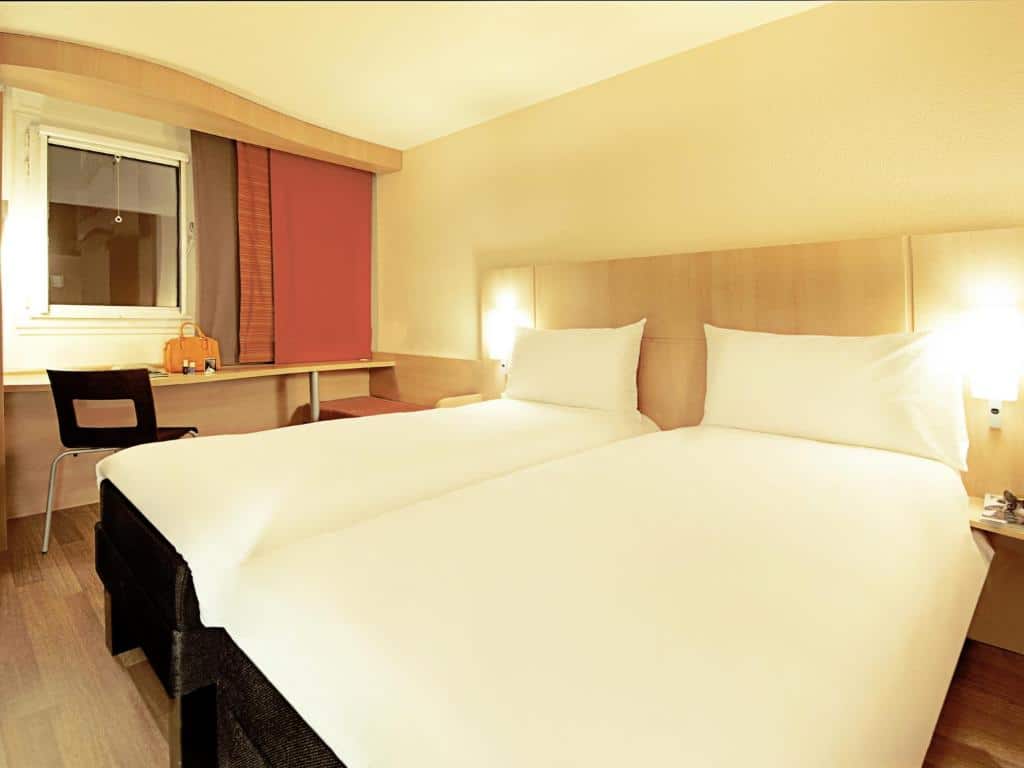 Quarto do Ibis Lisboa José Malhoa com duas camas de solteiro, uma secretária com uma cadeira e uma janela com estore