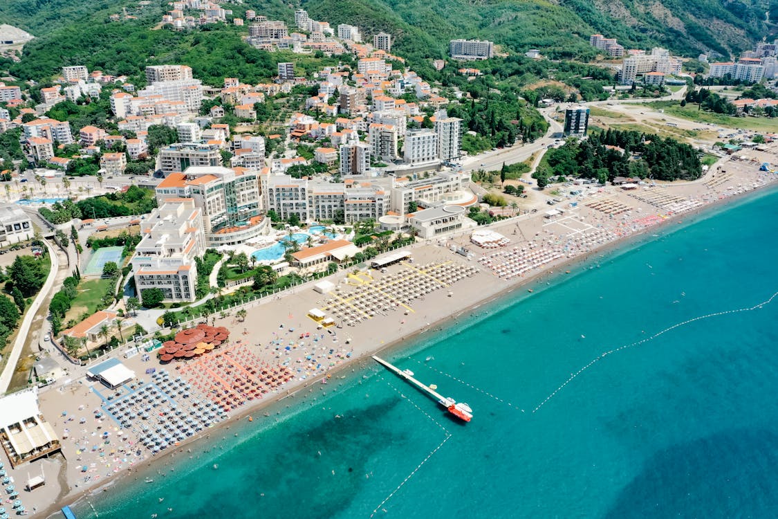 Vista aérea de uma praia em Budva, no Montenegro, com hotéis à beira-mar, chapéus de sol na areia e águas azul-turquesa cristalinas a estenderem-se até ao horizonte.