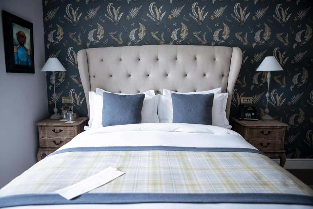 Uma cama bem feita com roupa de cama azul e branca entre duas mesas de cabeceira, contra uma parede com papel de parede estampado, no The Hayden Pub & Rooms.