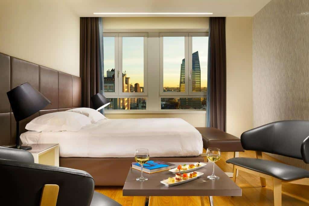 Quarto do UNAHOTELS Century Milano com janela com vista para a cidade, cama de casal com cabeceira em cabedal castanho, mesa com petiscos e vinho e duas poltronas acolchoadas