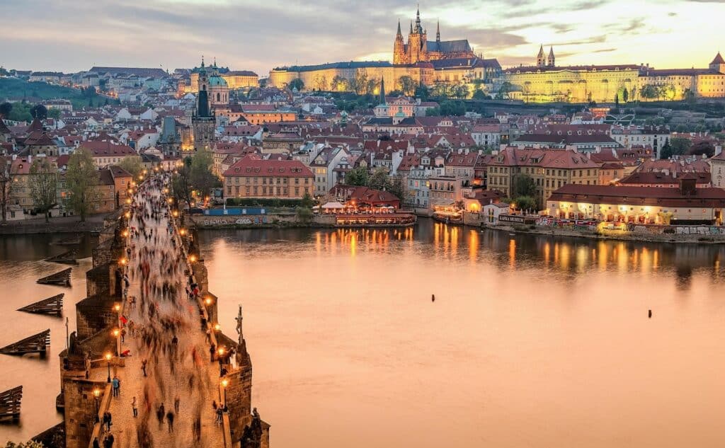 Praga, capital da República Checa, com uma ponte a atravessar o rio, com estátuas e pessoas a passar; do outro lado, a cidade e uma igreja no topo.
