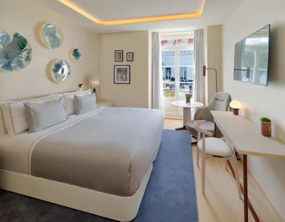 Quarto do The One Palácio da Anunciada com uma cama de casal, uma pequena varanda com cortinas e, em frente à cama, uma bancada com cadeira e uma televisão na parede; junto à varanda há uma poltrona estofada e um candeeiro de pé