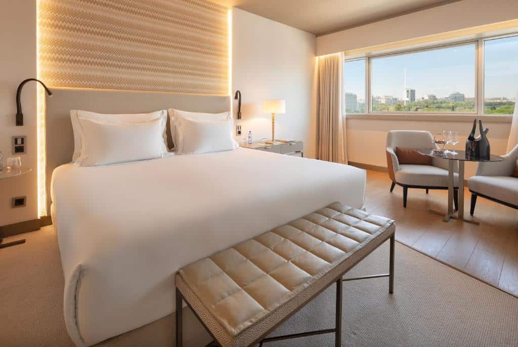 Quarto de hotel moderno com cama grande, banco, mesa pequena com duas cadeiras e janela com vista para a paisagem urbana.