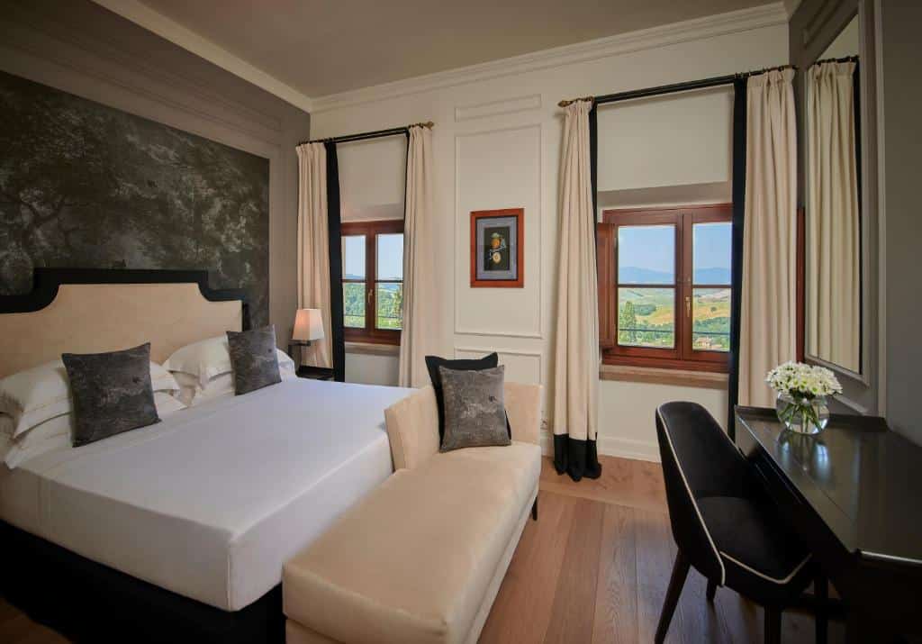 Quarto superior do Fonteverde Lifestyle &amp; Thermal Retreat, de 25 m², com cama de casal com almofadas cinzentas, sofá, mesa preta com um vaso de flores e cadeira estofada preta à frente, e duas janelas com vista sobre uma paisagem verde e o céu azul