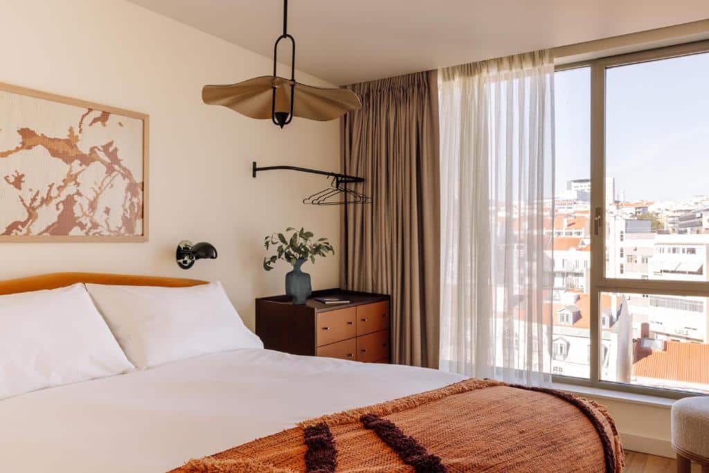 Quarto do Locke de Santa Joana com cama, mesa de cabeceira com uma planta, arte de parede, um bengaleiro e uma grande janela com vista para a cidade.