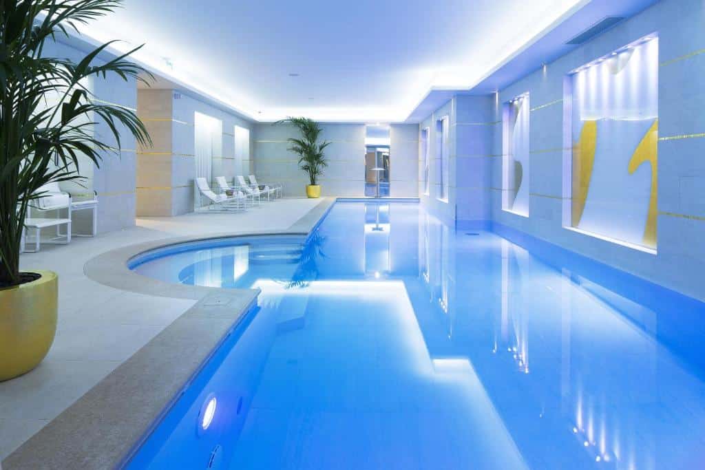 Piscina interior do Le Burgundy Paris com água azul, iluminação moderna, espreguiçadeiras, vasos de plantas e decorações abstratas nas paredes. Este é um dos hotéis de luxo em Paris.
