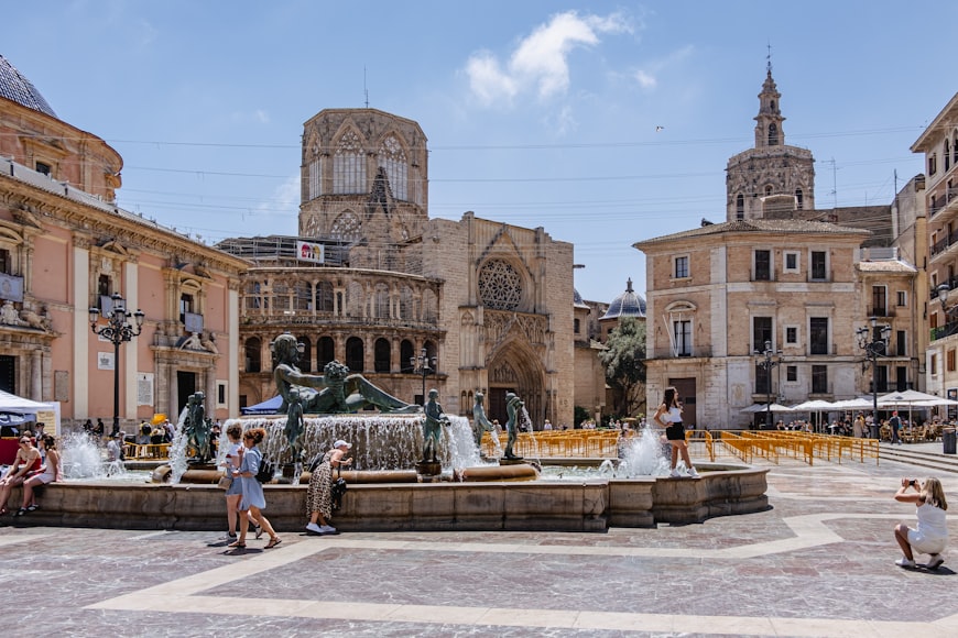 Praça com fonte e catedral gótica no centro histórico de Valência, com pessoas a caminhar e a aproveitar um dia soalheiro.