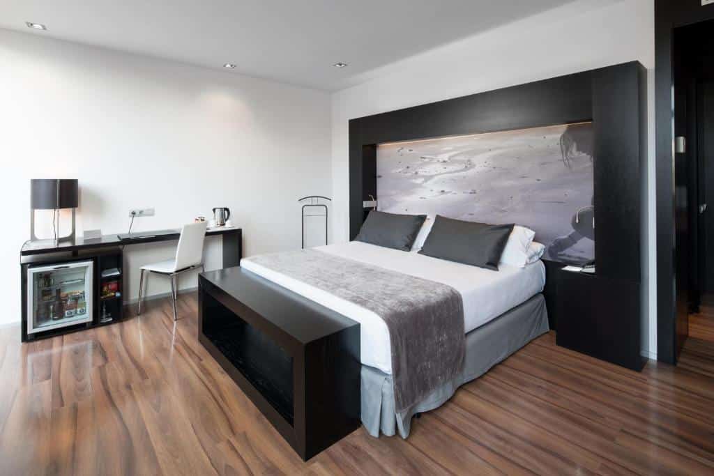 Quarto do Catalonia Catedral, uma das recomendações de hotéis no Bairro Gótico em Barcelona. A cama de casal tem mesas de cabeceira de ambos os lados, um móvel de madeira aos pés e uma secretária com candeeiro e frigobar.