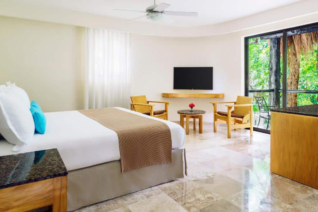 Quarto do The Reef Playacar Beach Resort & Spa com cama de casal ao centro, mesa de cabeceira e vista para área verde tropical pela varanda.