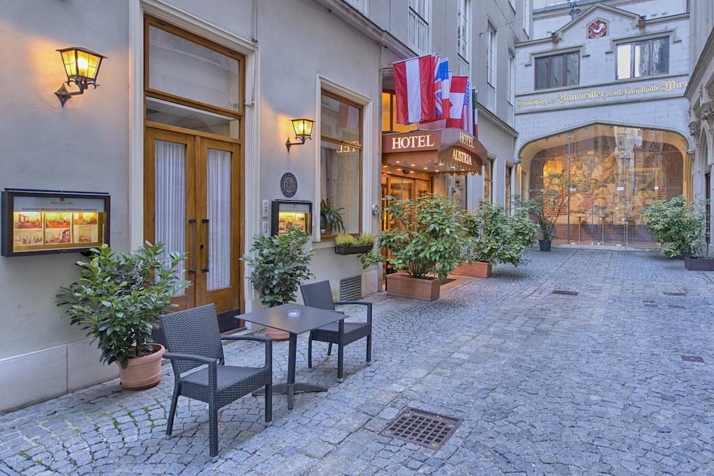 Esplanada com duas cadeiras e uma mesa em frente à entrada do Hotel Austria – Wien, numa rua de calçada com vasos de plantas.