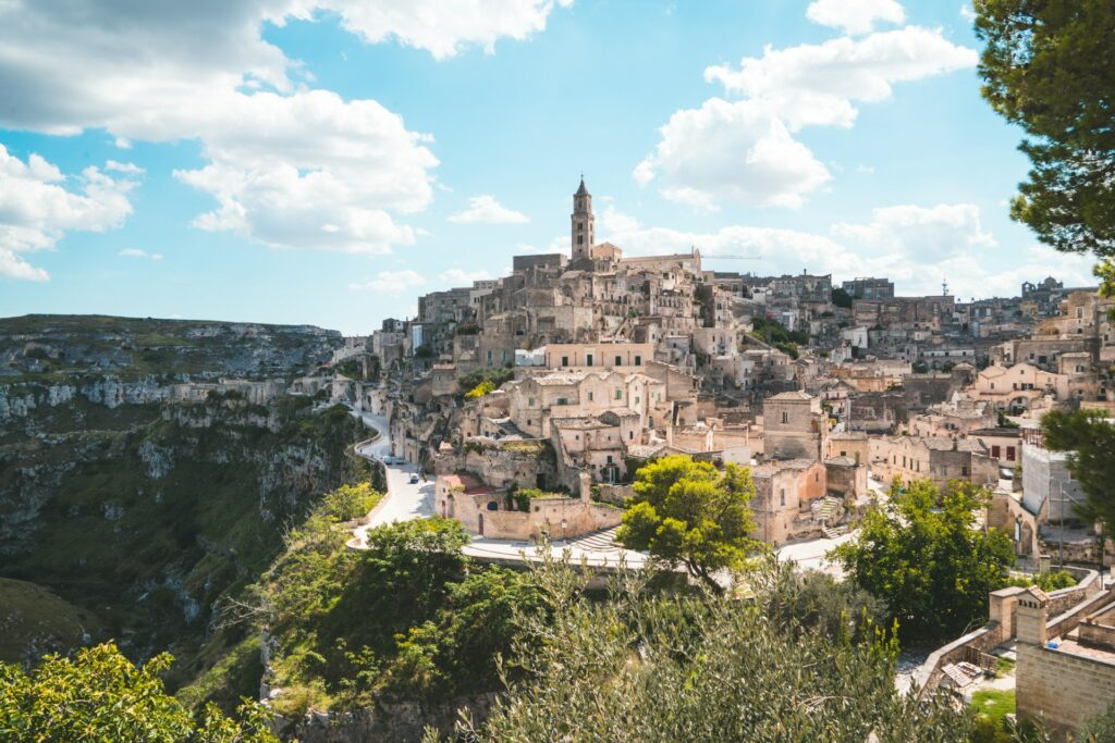 Vista de Matera, Itália, famosa pelas habitações escavadas na rocha, conhecidas como "Sassi". A cidade estende-se ao longo de um penhasco com vista para um vale profundo. Uma igreja com uma torre alta destaca-se no centro, sob um céu azul com nuvens.
