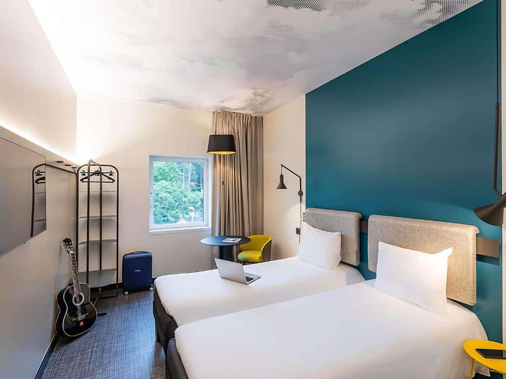 Quarto do Hotel Ibis Lisboa Alfragide com duas camas de solteiro, parede com detalhes em azul-petróleo, candeeiros de parede e mesa redonda.