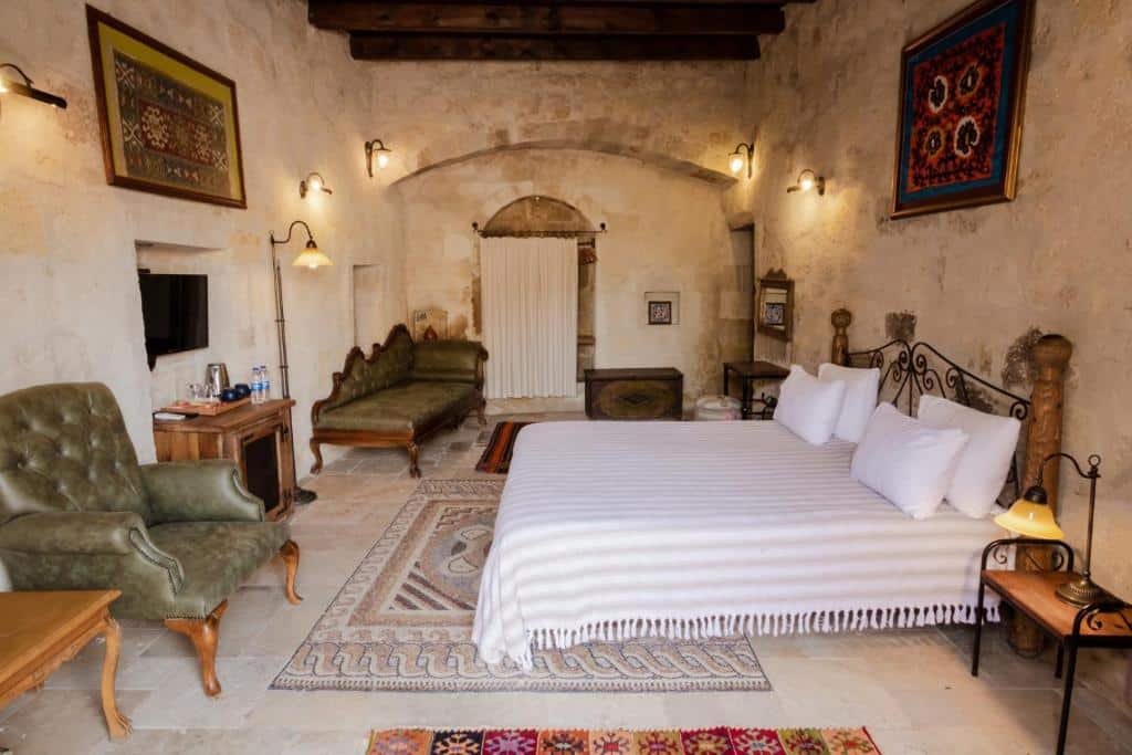 Quarto espaçoso do Sultan Cave Suites, um dos hotéis da Capadócia, com paredes de pedra, cama grande, poltrona, sofá, tapetes, mobiliário de madeira e arte na parede.