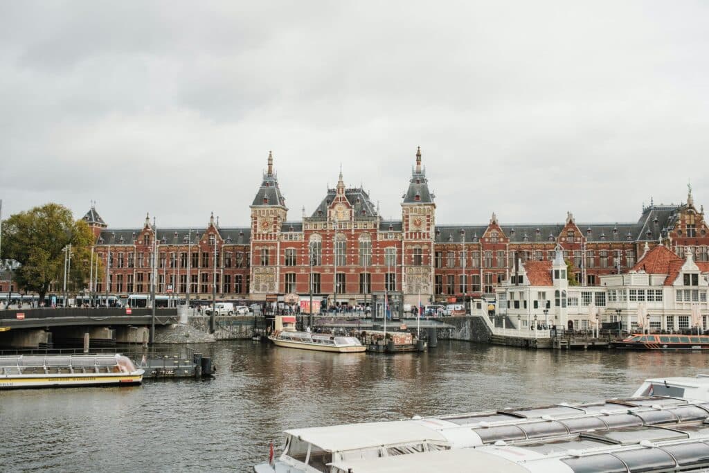 Grande edifício de tijolo vermelho com torres junto a um canal, barcos atracados ao longo da margem e céu nublado.