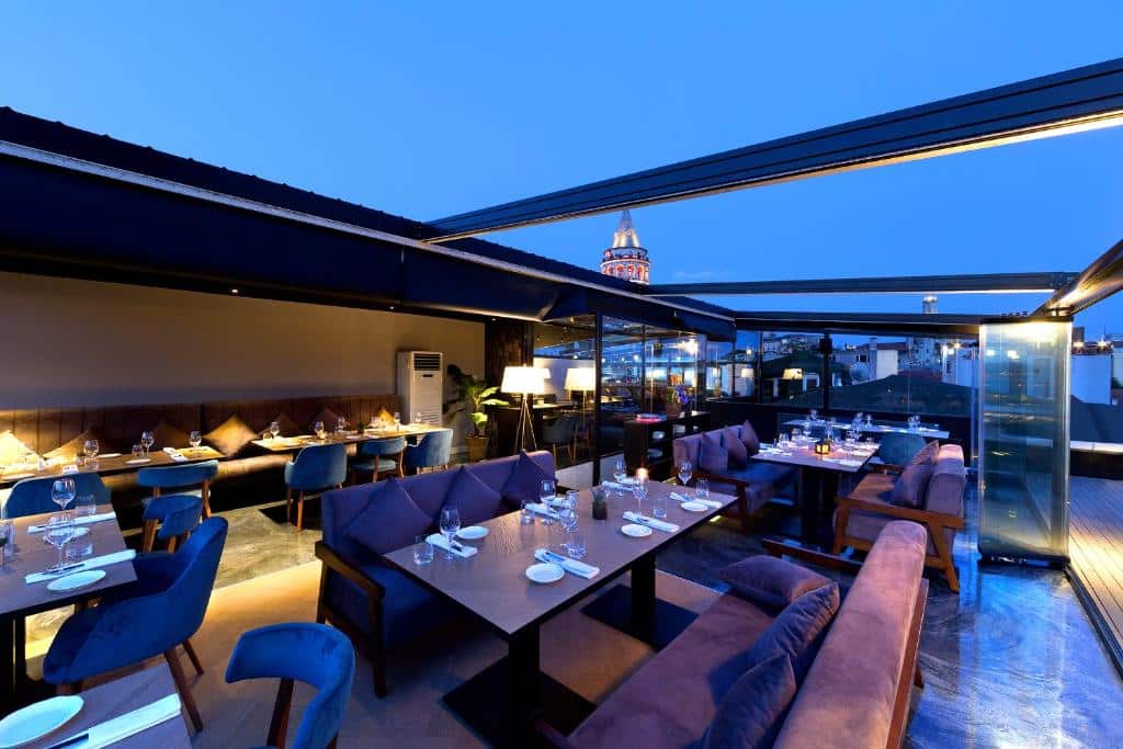 Restaurante do Walton Hotels no terraço ao anoitecer, com mesas postas, assentos almofadados e vistas da cidade através de painéis de vidro abertos.