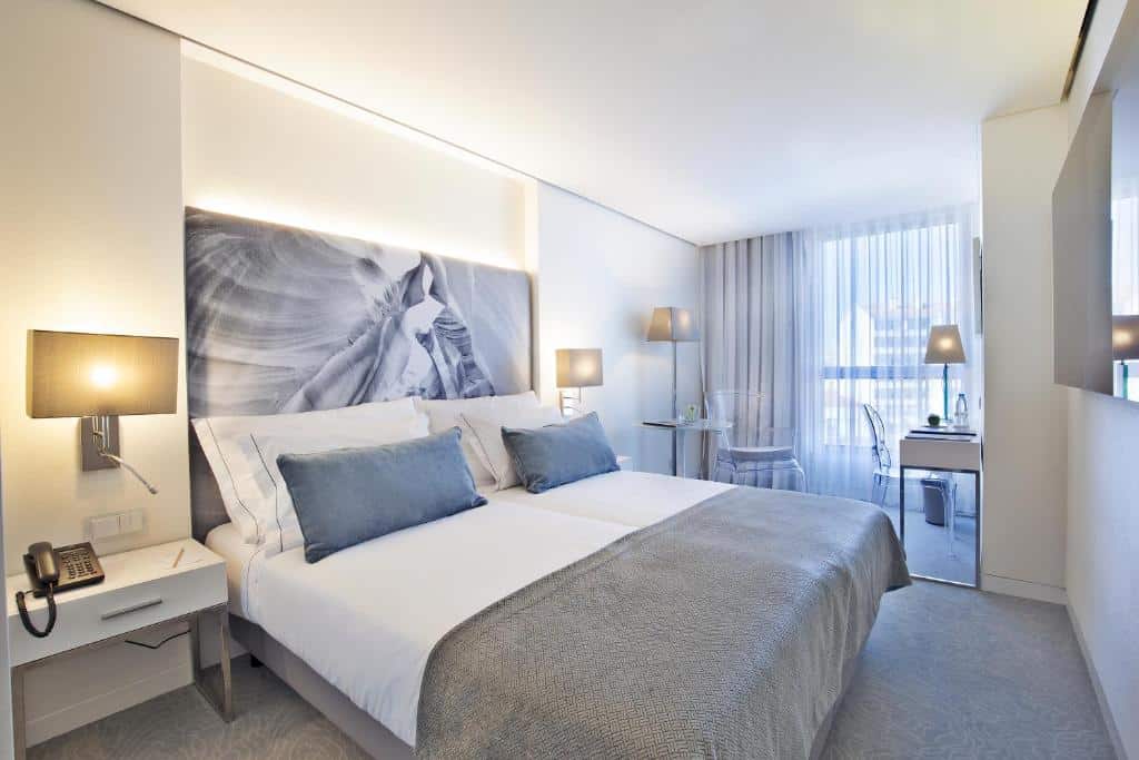 Quarto do Hotel White Lisboa com cama de casal, uma janela ampla com cortinas brancas e, junto dela, uma secretária com cadeira; em frente à cama, uma televisão na parede