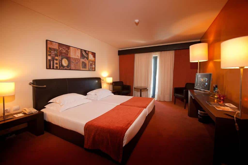 Quarto do Vila Gale Santa Cruz com cama de casal do lado esquerdo da imagem em cada lado da cama uma cômoda com luminária em frente a cama uma cômoda com TV. Representa hotéis all inclusive em Portugal.