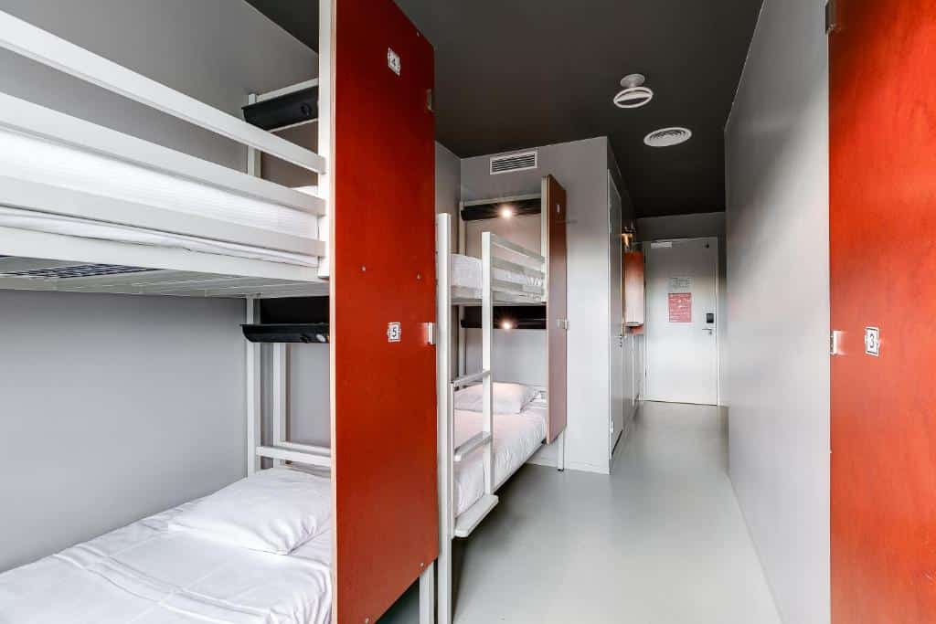 Quarto do ClinkNOORD Hostel, que ilustra onde ficar em Amesterdão: quarto estreito com dois beliches, roupa de cama branca, cacifos vermelhos e uma porta ao fundo do corredor.