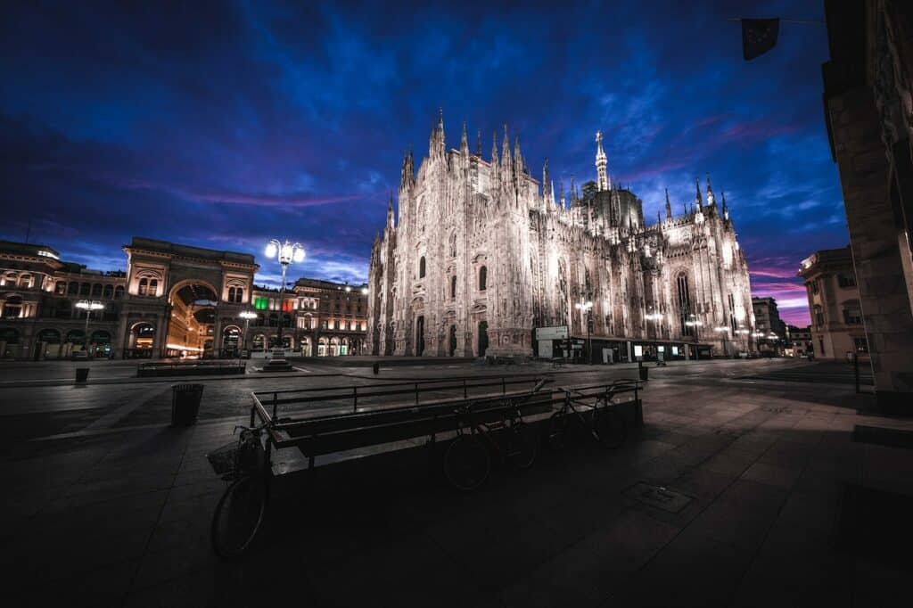 Catedral de Milão em estilo gótico iluminada à noite, com a Praça Duomo vazia e outros edifícios históricos em redor