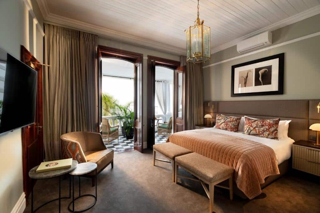 Quarto no The Winchester Hotel com cama grande, poltrona, TV e varanda com assentos ao ar livre e vegetação lá fora.