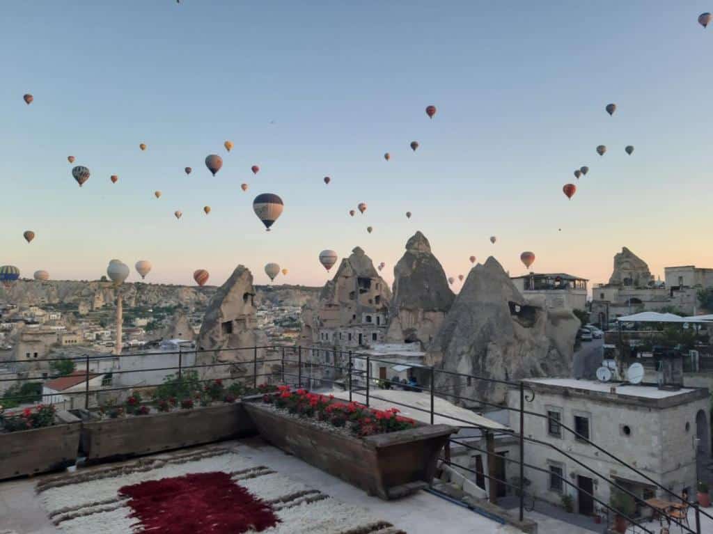Balões de ar quente sobre formações rochosas e edifícios ao nascer do sol, vistos de um terraço com floreiras e tapetes, no Artemis Cave Suites & Spa.