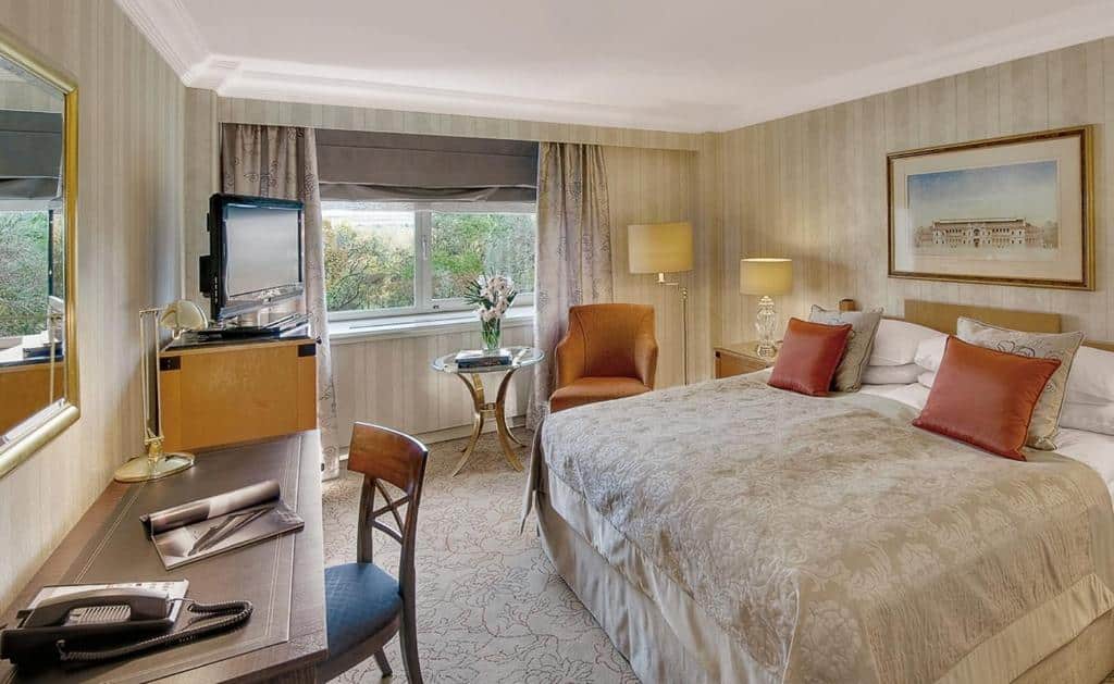 Quarto no InterContinental Wien, com cama de casal, secretária, cadeira, TV, poltrona, mesa lateral com flores e uma janela com cortinados.