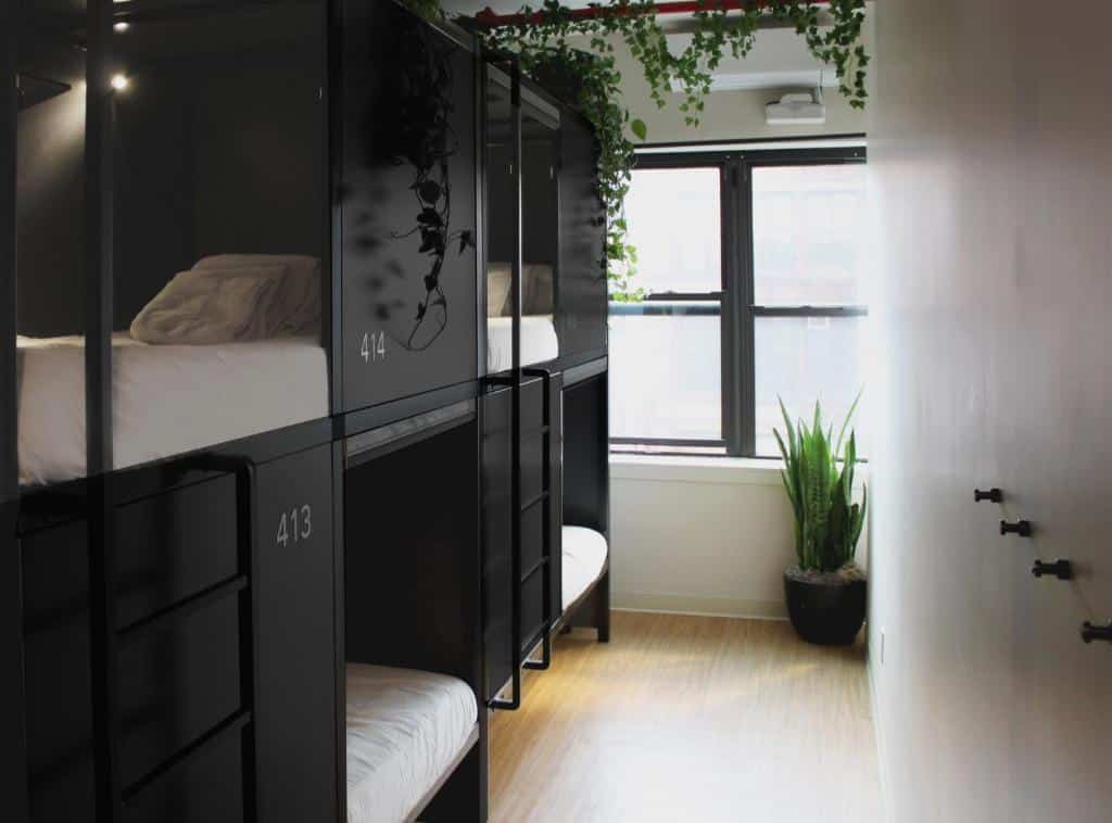 Quarto estreito com quatro beliches pretos, numerados 413 e 414, roupa de cama branca, plantas e uma janela ao fundo.