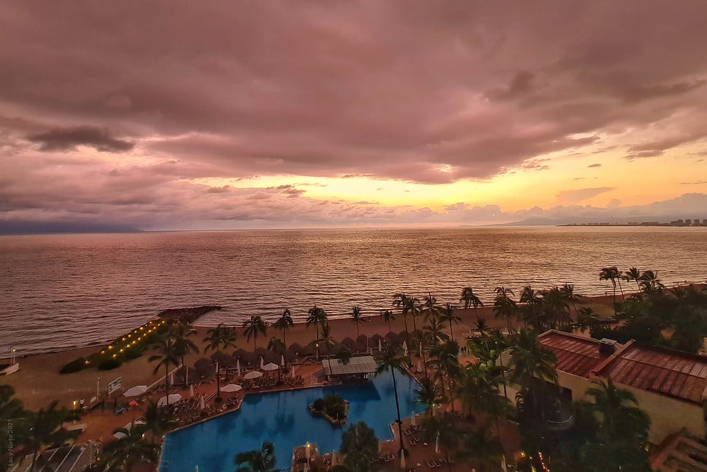 Vista de um resort à beira-mar ao pôr do sol com piscina, palmeiras e céu nublado com vista para o oceano.