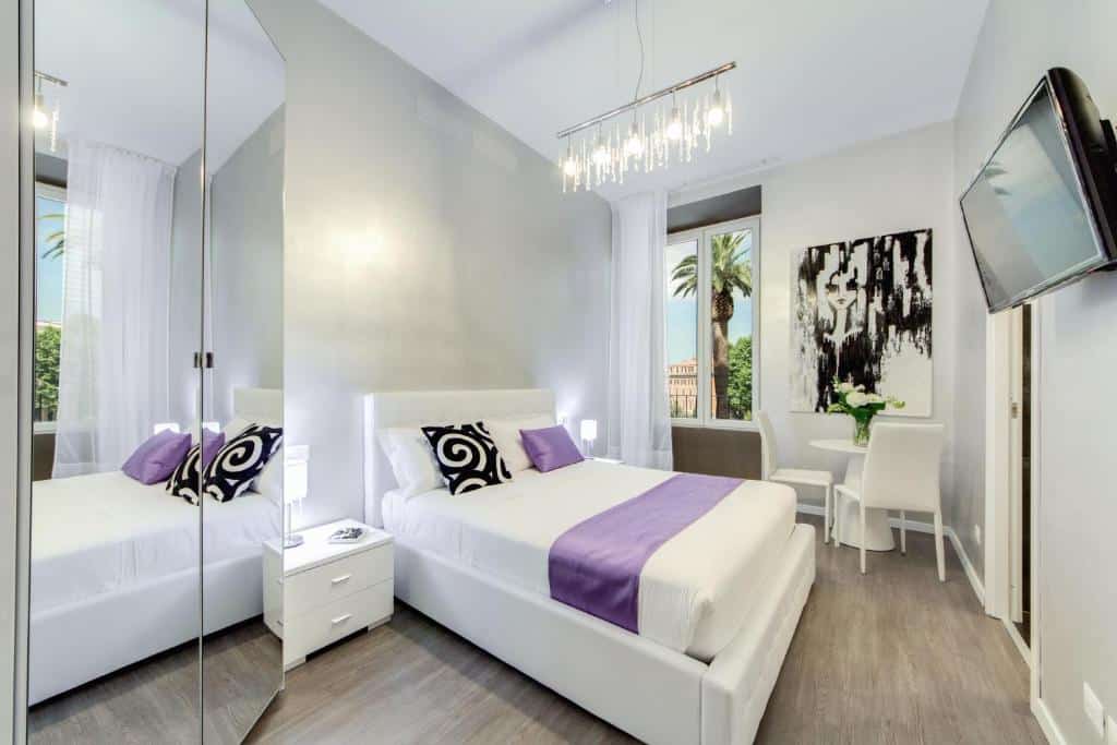 quarto do B&amp;B Colosseo Panoramic Rooms, com cama de casal, roupeiro espelhado, televisão em frente, quadro na parede, janela com cortinas, um lustre comprido por cima e mesa de cabeceira com candeeiro de cada lado, além de uma mesa e duas cadeiras