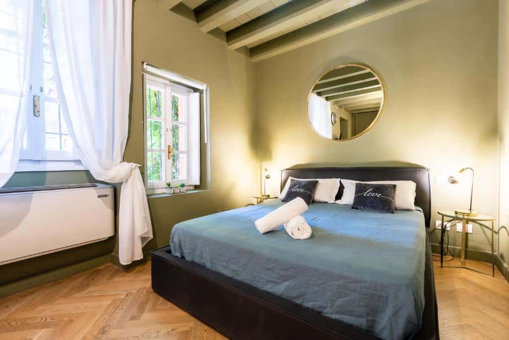 Quarto do Alzaia Naviglio Grande com duas janelas com cortinas, uma cama de casal, um espelho redondo por cima da cama e mesas de cabeceira com candeeiros nas laterais