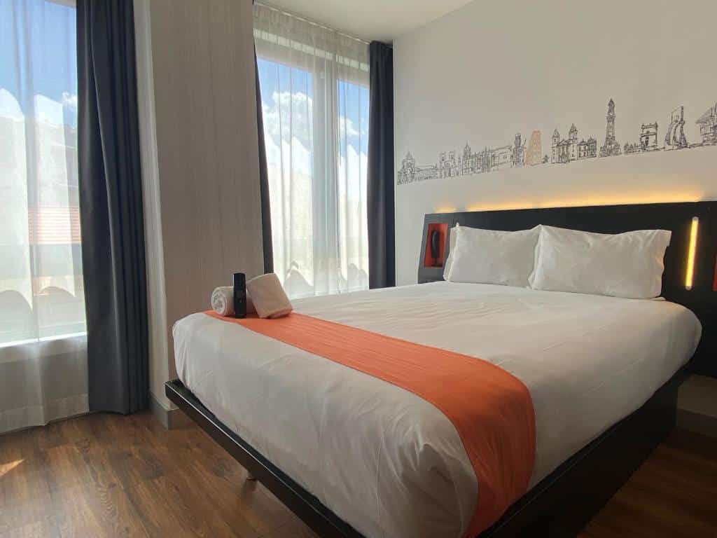Quarto do easyHotel Lisbon com uma cama de casal e, do lado esquerdo, duas janelas com cortinas brancas; o chão imita madeira.