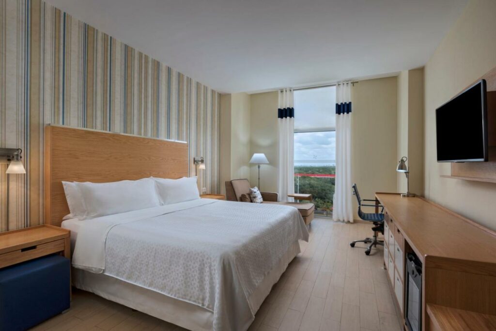 Quarto no Four Points by Sheraton Cancun Centro com varanda, mesa com cadeira, televisão, poltrona, cama e duas mesas de cabeceira com candeeiros