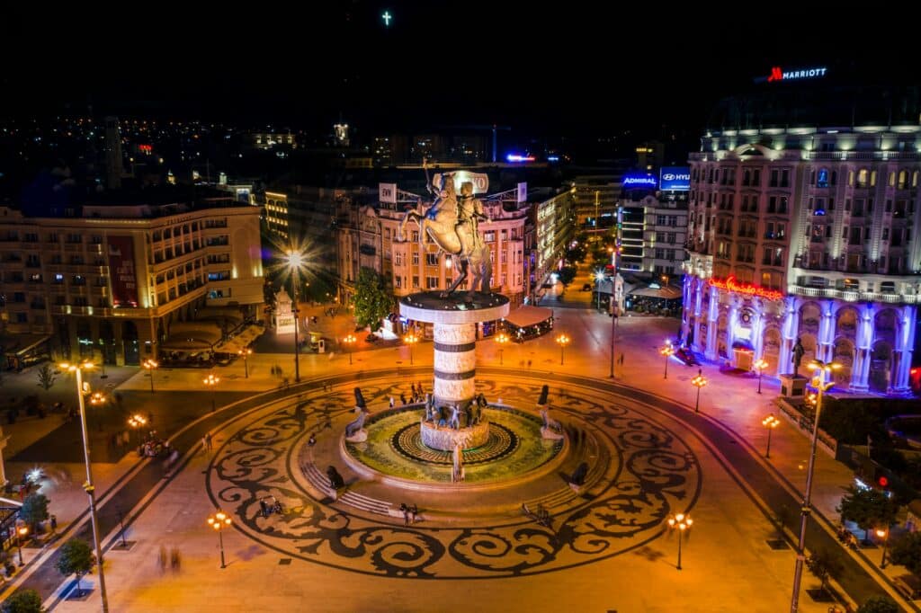 Vista aérea noturna de uma praça em Skopje, a capital da Macedónia do Norte, um dos destinos invulgares da Europa. O foco é uma grande estátua no centro da praça, iluminada e rodeada por padrões circulares no chão. As ruas em redor estão repletas de luzes, e edifícios modernos e históricos iluminados ladeiam a cena.
