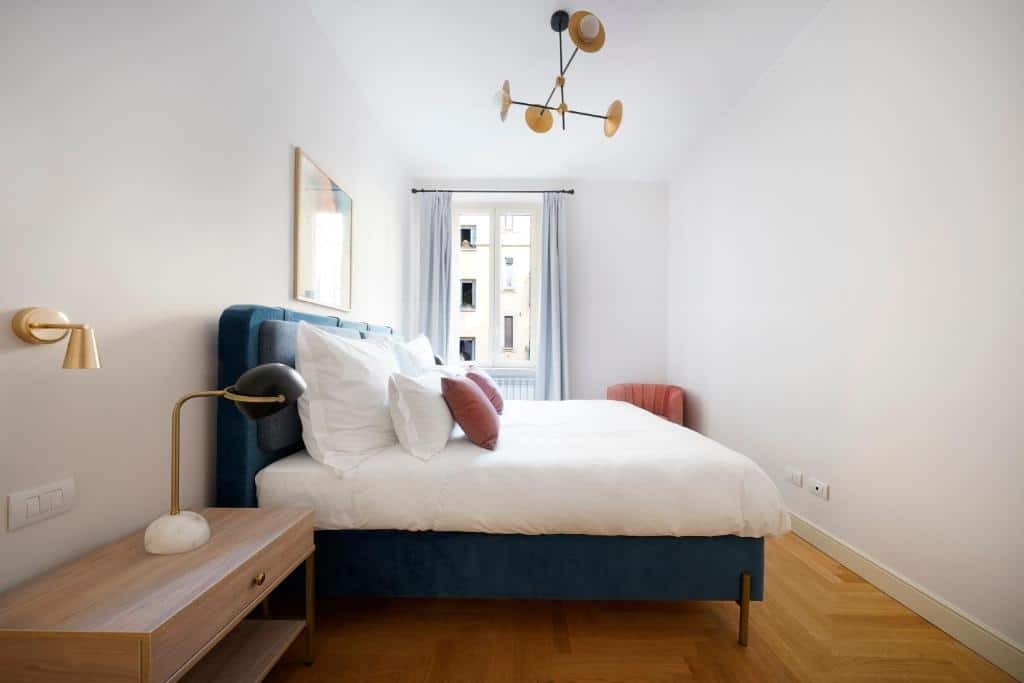 Quarto do numa I Loreto Apartments com uma cama de casal, chão em imitação de madeira, uma janela com cortinas, uma mesa de cabeceira com um candeeiro e um lustre com várias lâmpadas