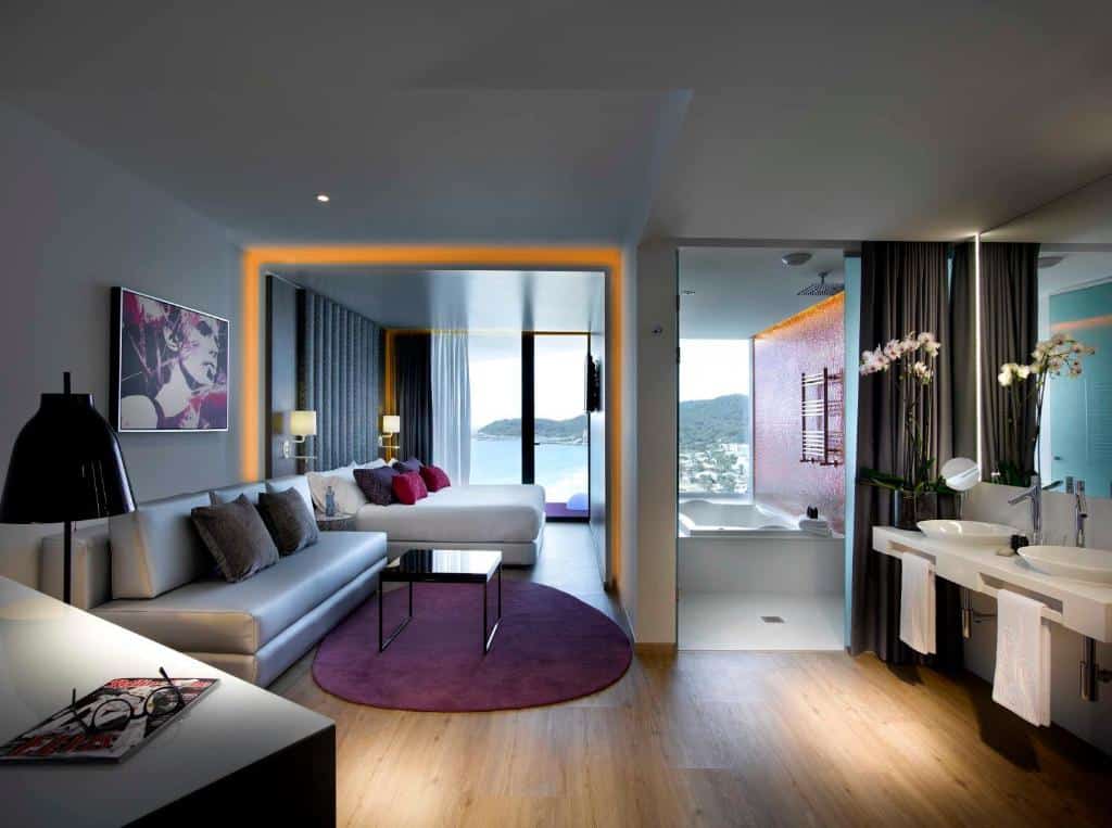 Suíte moderna do Hard Rock Hotel Ibiza com zona de estar, cama, varanda, vista para o mar e casa de banho aberta com lavatórios duplos e chuveiro.