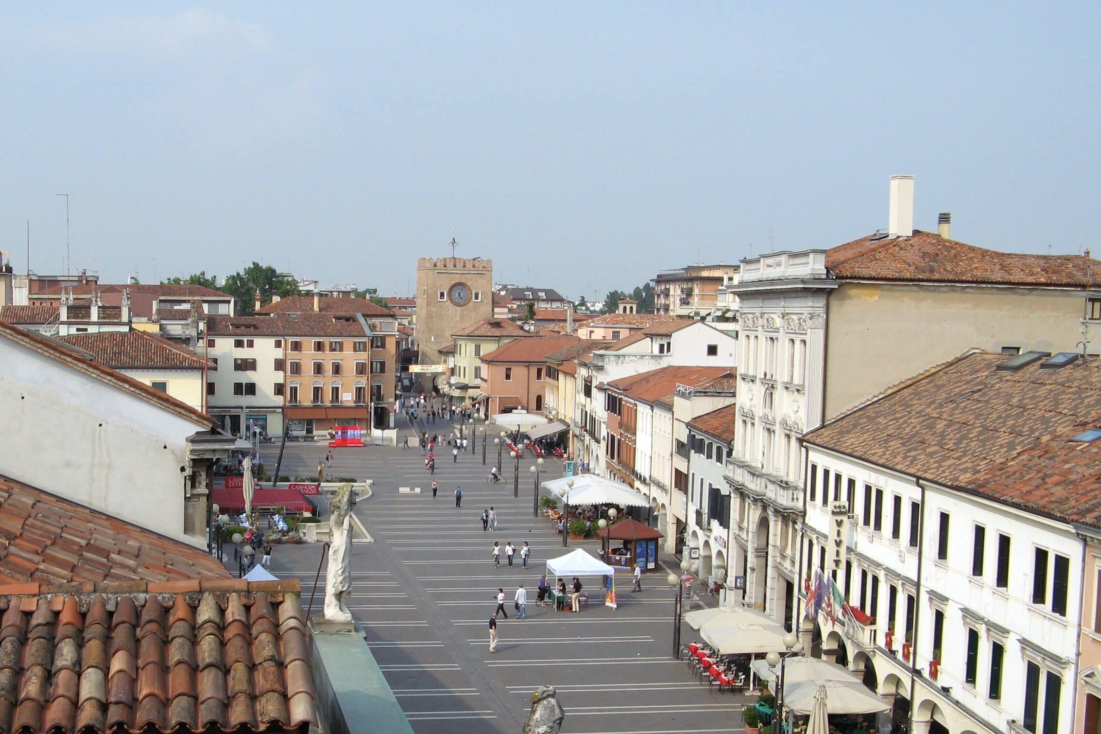 Praça no centro histórico de uma cidade italiana, com edifícios coloridos, igreja com torre do relógio e transeuntes na escadaria