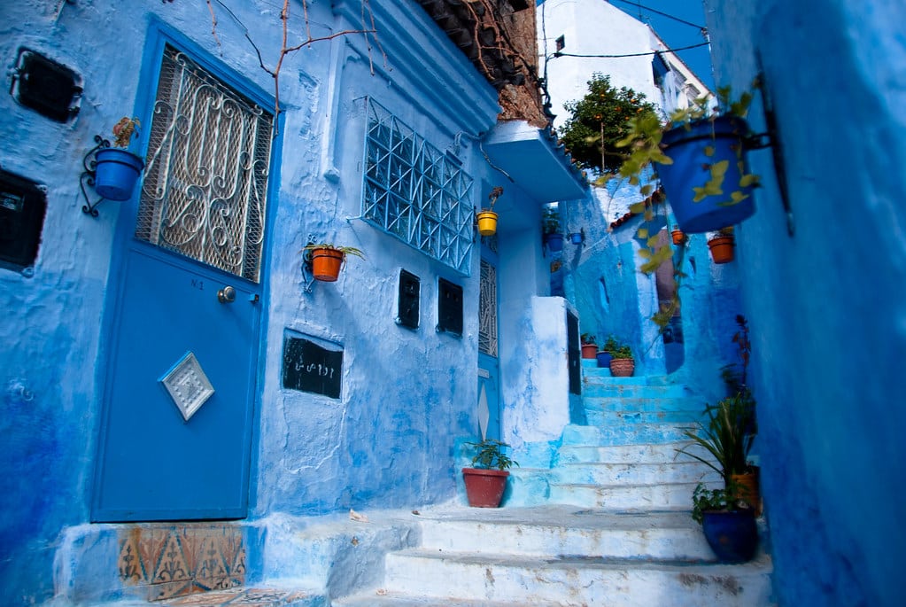 Um beco estreito com edifícios pintados de azul e plantas em vasos nas escadas, em Chefchaouen, Marrocos, sob um céu azul claro.