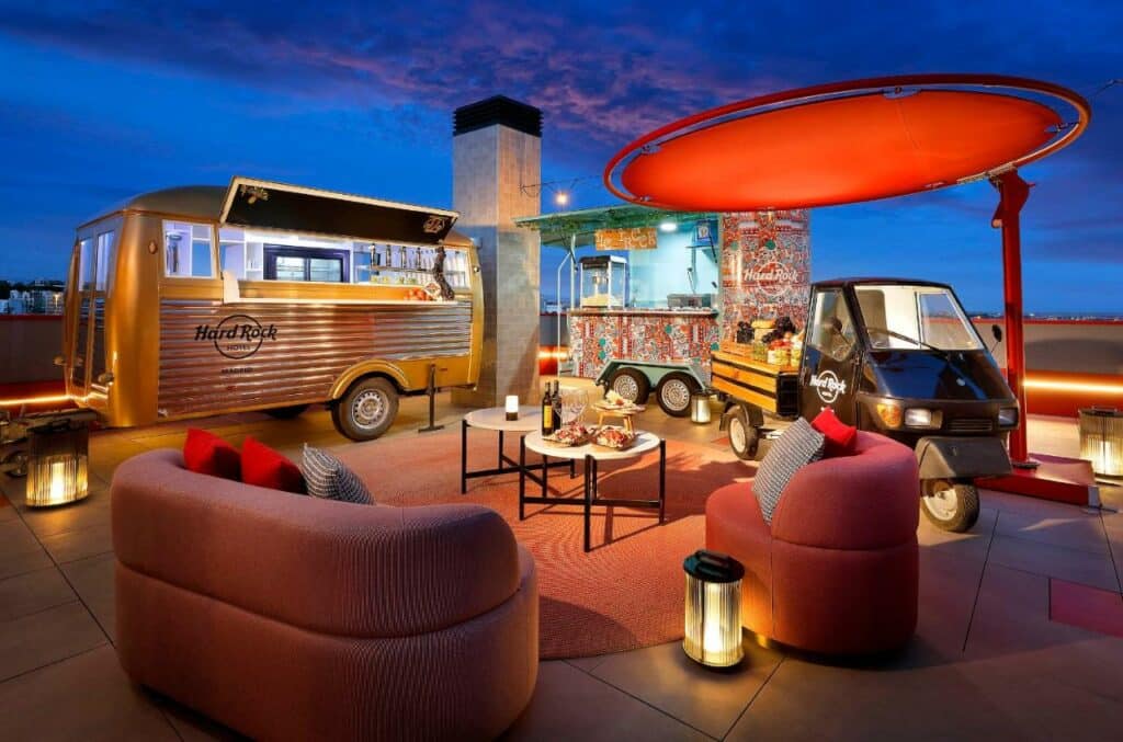 Roulottes de bebidas e comidas no terraço do Hard Rock Hotel Madrid. Duas poltronas estão viradas para uma mesa ao centro com vários itens.