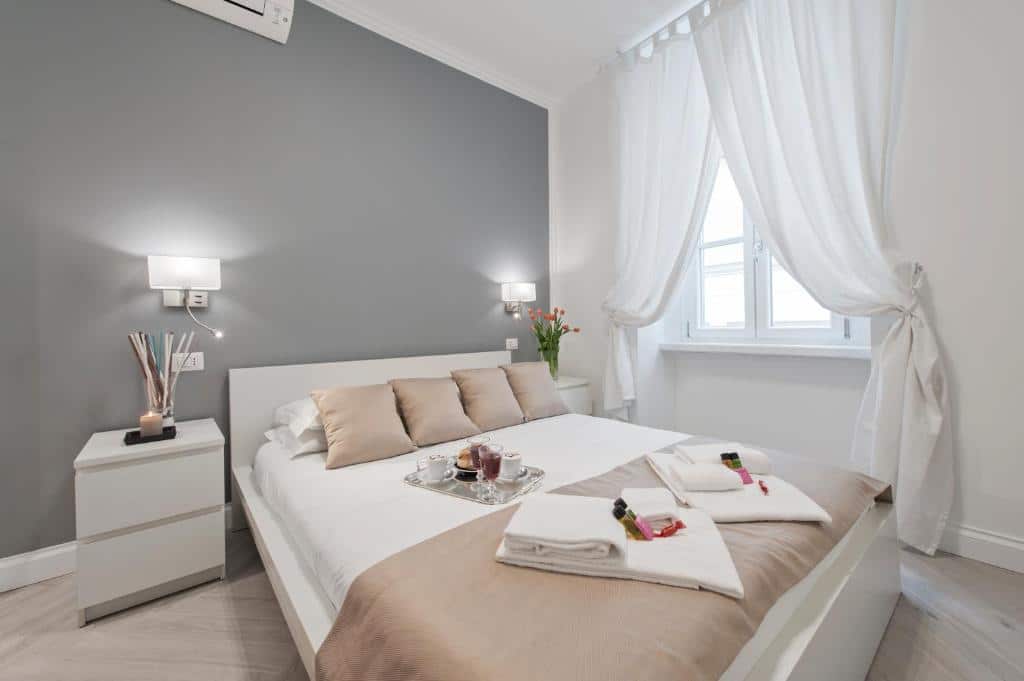 quarto do Trevi Apartment, um alojamento local em Roma, com cama grande e pormenores em rosé, cinza e branco, uma janela com cortinas leves, candeeiro e mesa de cada lado da cabeceira e ar condicionado